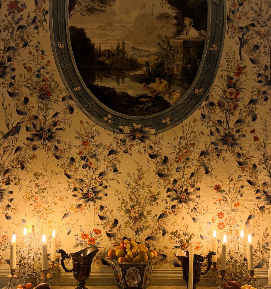 Traditional floral wallpaper with ribbon and bird motifs above a marble mantel lit by candles