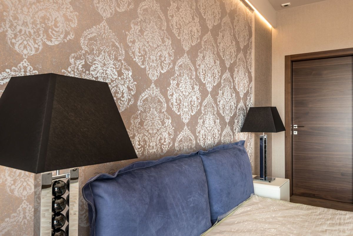Bedroom with a metallic damask wallpaper feature wall behind a blue upholstered bed and black table lamps