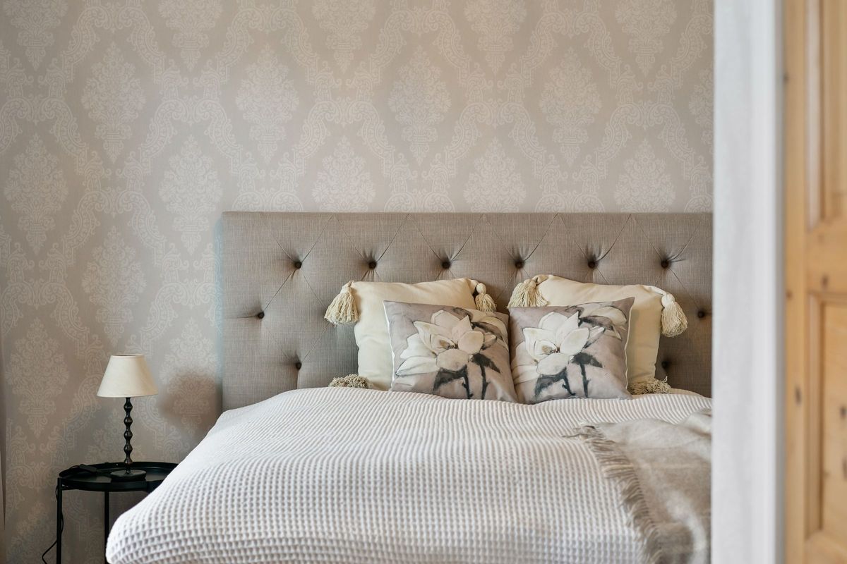 Soft neutral damask wallpaper behind a tufted linen headboard and floral cushions